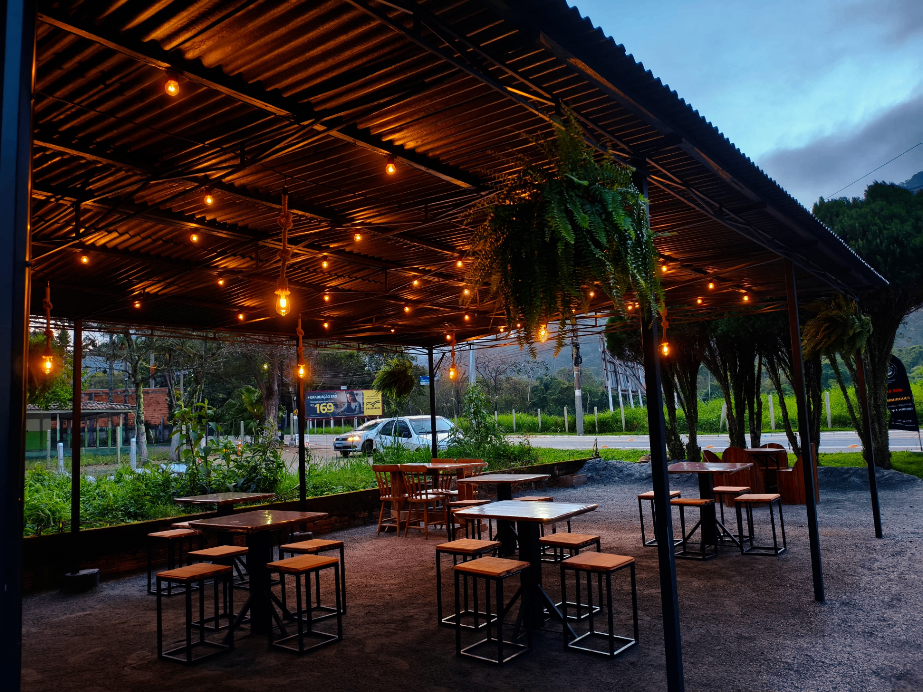 Espaço coberto do Estaleirinho Burger com lâmpadas pendentes, samambaias e mesas industriais